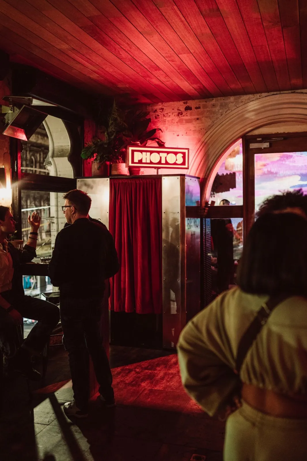 A stainless-steel photo booth with a red velvet curtain and a lit PHOTOS sign in a busy bar