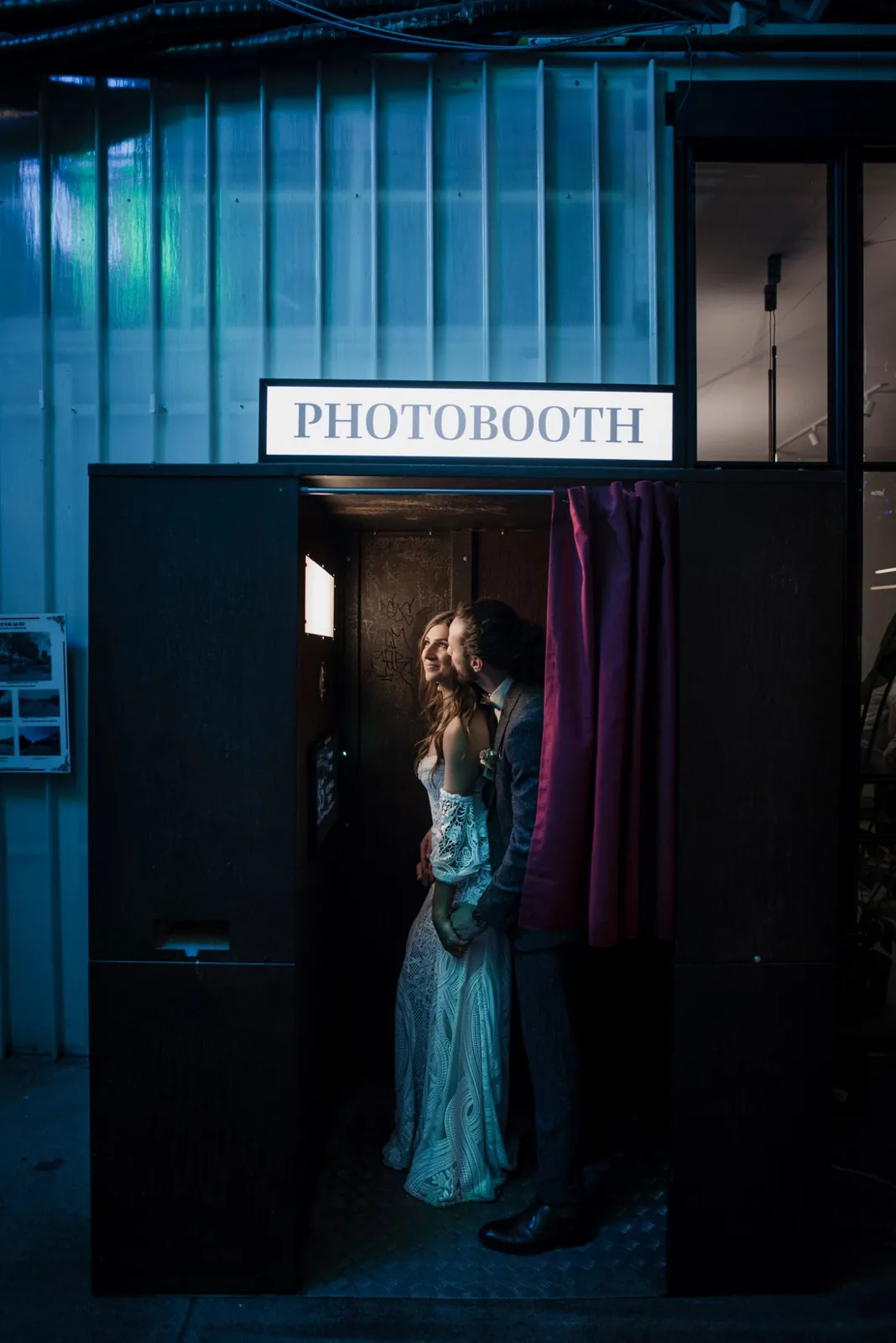 A bride and groom inside a black photo booth under a lit PHOTOBOOTH sign