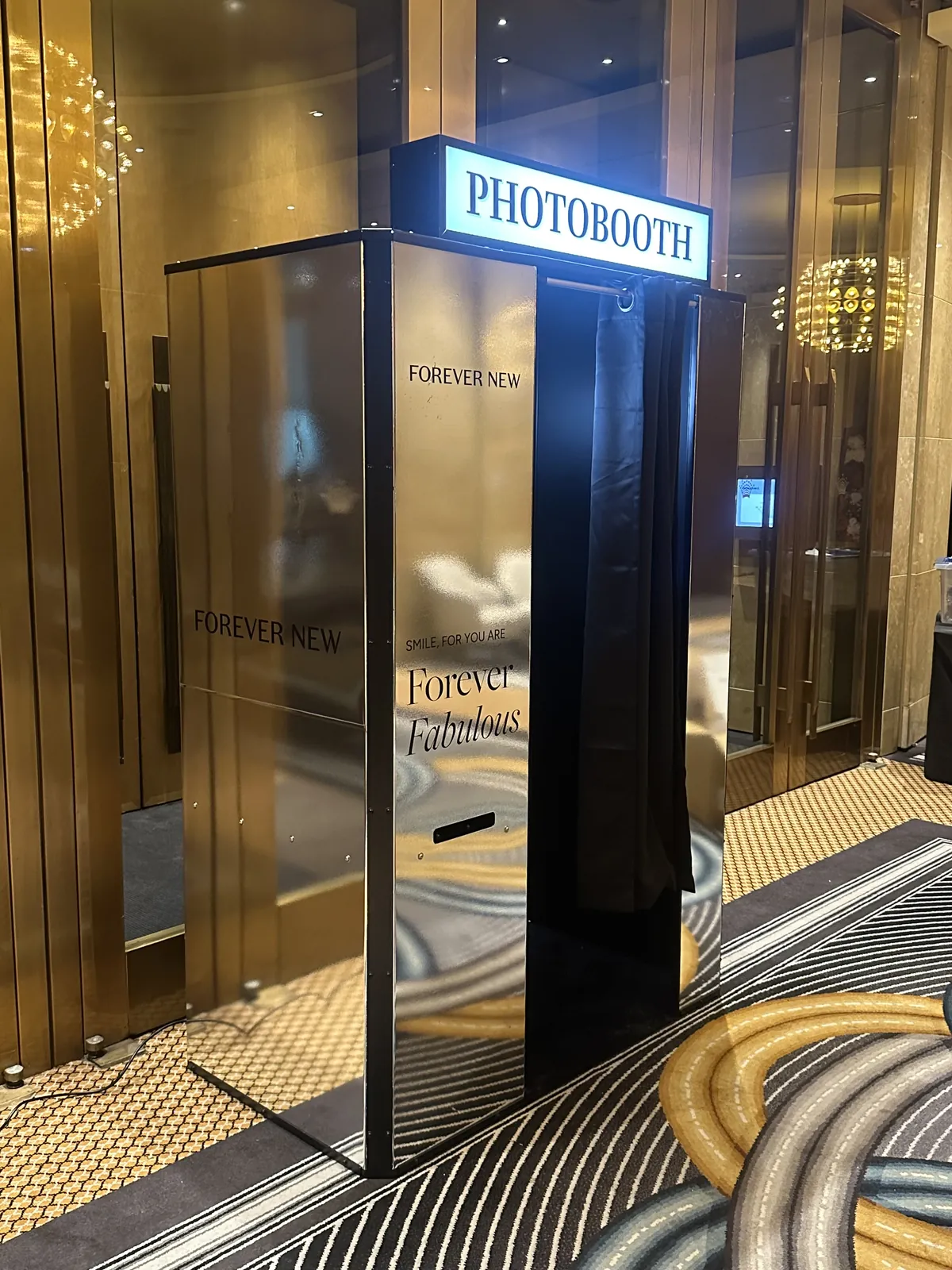 A mirror-finish photo booth wrapped in Forever New branding under a lit PHOTOBOOTH sign